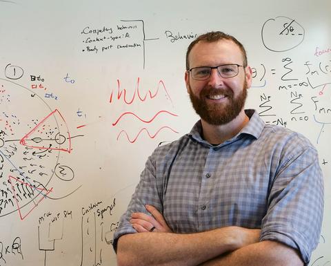 Man with beard and glasses wearing a plaid shirt standing in front of a white board with writing on it