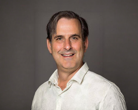 Man with dark hair wearing a white shirt and smiling in front of a dark gray background
