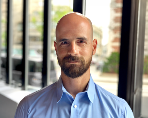 Man with beard wearing a blue shirt standing in front of windows