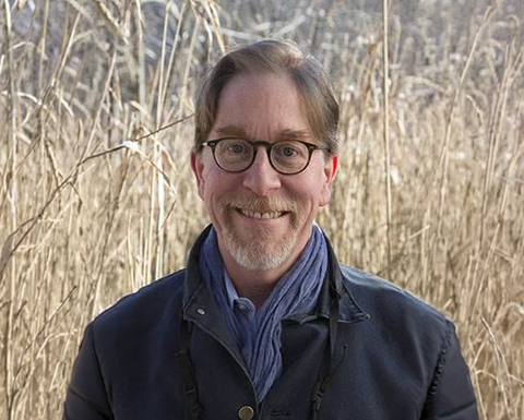 Man wearing glasses, a black jacket and scarf standing in a field 