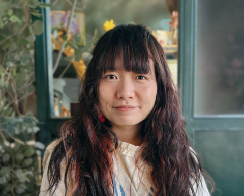 Woman with long dark hair and bangs sitting in front of plants and windows