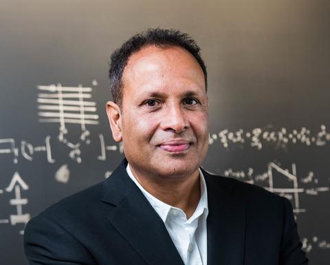 Man with dark hair wearing a black suit and standing in front of a chalk board with writing