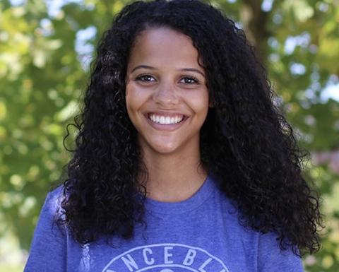 Maya Woolfolk with long curly black hair, wearing a blue shirt with "dance blue" on it, in front of leafy green trees