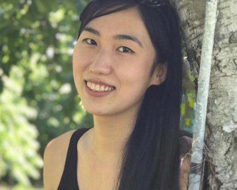 Zhe He with long dark hair, wearing a black top with sunglasses on her head, leaning against a tree